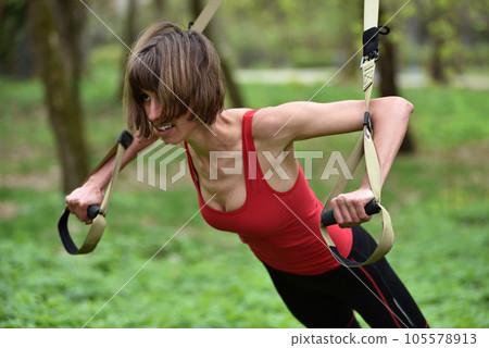 Young woman does suspension training with fitness straps in the outdoors Young woman does suspension training with fitness straps in the outdoors 105578913