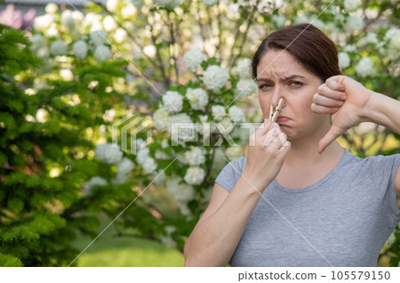 A woman suffers from a clothespin on her nose and shows a thumbs down on a walk in a flowering park. Allergy. A woman suffers from a clothespin on her nose and shows a thumbs down on a walk in a flowering park. Allergy. 105579150