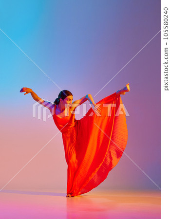 Grace and tenderness. Young beautiful woman in long elegant dress dancing against gradient multicolor background in neon light Grace and tenderness. Young beautiful woman in long elegant dress dancing against gradient multicolor background in neon light 105580240