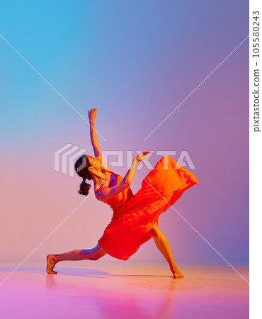 Dynamic image of artistic young woman dancing in elegant red dress against gradient multicolor background in neon light 105580243
