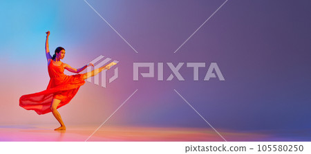 Elegant, talented young woman in red long red dress dancing against gradient multicolor background in neon light 105580250