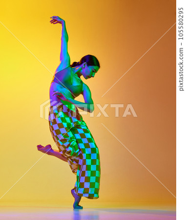 Full-length image of young artistic, expressive woman in stylish costume dancing against gradient yellow orange background in neon light 105580295