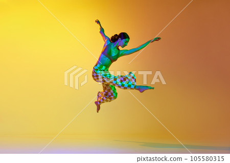 Young woman, professional contemporary dancer in motion, in stylish clothes against gradient yellow orange background in neon light 105580315