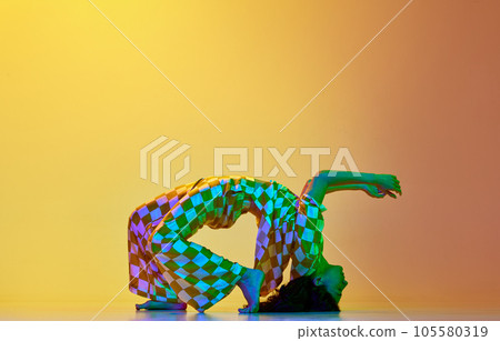 Dynamic image of flexible, artistic young woman dancing against gradient yellow orange background in neon light 105580319