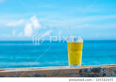 Big glass drink plastic straw green tea looks like yellow beer or kombucha blue sea white clouds background water drops condensed 105582035