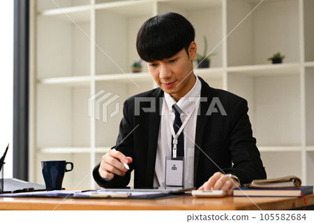 Asian male manager wearing black suit hand filling paper business document, signing contract at office desk. Asian male manager wearing black suit hand filling paper business document, signing contract at office desk. 105582684