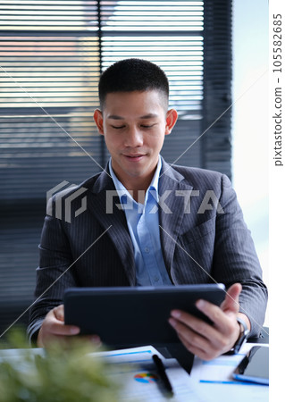 Millennial Asian male investor analyzing sales statistics data on digital tablet. Millennial Asian male investor analyzing sales statistics data on digital tablet. 105582685