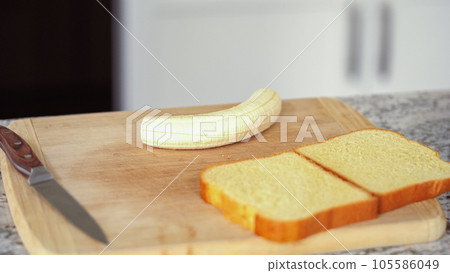 In a modern white kitchen, buttered banana and brioche bread are being prepared for making a delicious grilled peanut butter banana sandwich. In a modern white kitchen, buttered banana and brioche bread are being prepared for making a delicious grilled peanut butter banana sandwich. 105586049