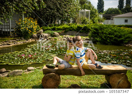 Two brother boys sitting on a bench in a park near peaceful lake with water lily Nymphaeum. Kids relaxing on nature on hot summer day Two brother boys sitting on a bench in a park near peaceful lake with water lily Nymphaeum. Kids relaxing on nature on hot summer day 105587347