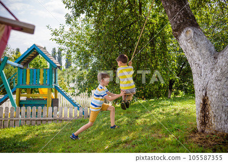 Two adorable happy little boys is having fun on a rope swing which he has found while having rest outside city. Active leisure time with children Two adorable happy little boys is having fun on a rope swing which he has found while having rest outside city. Active leisure time with children 105587355