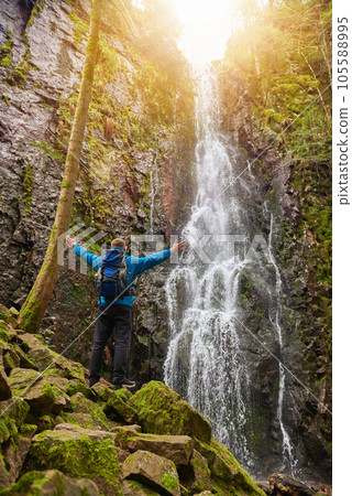 Tourist attraction of Germany - falls of Burgbach Waterfall near Schapbach, Black Forest, Baden-Wurttemberg, Germany. Man hiker in blue jacket standing on stone and looks at flow of falling water Tourist attraction of Germany - falls of Burgbach Waterfall near Schapbach, Black Forest, Baden-Wurttemberg, Germany. Man hiker in blue jacket standing on stone and looks at flow of falling water 105588995