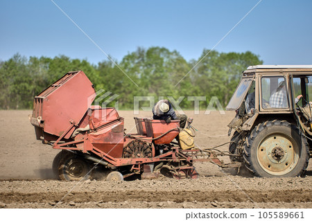 Planting potatoes with potato planter hitched to wheeled agricultural tractor. Planting potatoes with potato planter hitched to wheeled agricultural tractor. 105589661