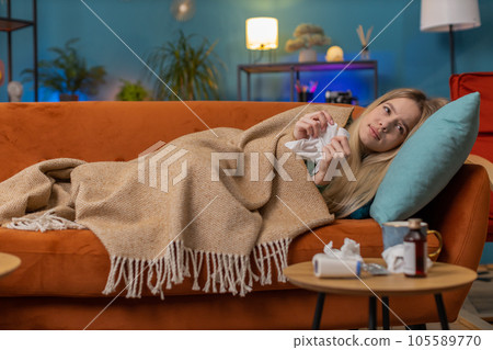 Sick ill young woman suffering from cold, allergy, lying on home sofa sneezes wipes snot into napkin Sick ill young woman suffering from cold, allergy, lying on home sofa sneezes wipes snot into napkin 105589770