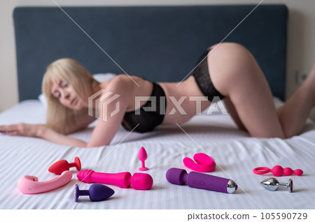 Blonde in lingerie lies on the bed near the collection of vibrators.  105590729