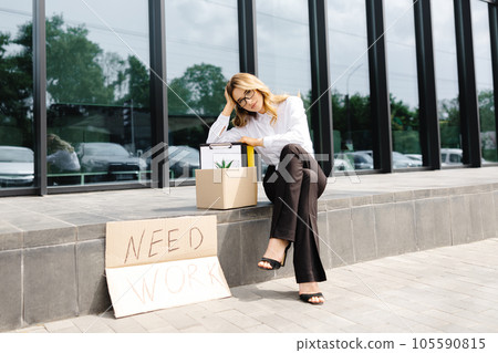Woman sitting outdoor with cardboard banner with the slogan Need work next to box of stuff. Beautiful female protester at strike against unemployment 105590815