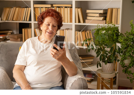 Portrait of a modern elderly woman who sits in an armchair against the background of a bookcase with books and a flower, looks at the phone and smiles during the day in the living room Portrait of a modern elderly woman who sits in an armchair against the background of a bookcase with books and a flower, looks at the phone and smiles during the day in the living room 105591834