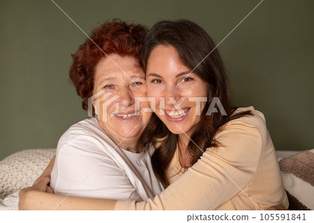 Close-up portrait of a cheerful grandmother and granddaughter sitting on the sofa during the day in the living room and hugging each other tightly with their arms and looking at the camera Close-up portrait of a cheerful grandmother and granddaughter sitting on the sofa during the day in the living room and hugging each other tightly with their arms and looking at the camera 105591841