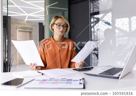 Serious and pensive business woman behind paper work inside office, female financier worker thinks about contracts and reports with charts and graphs, blonde successful woman uses laptop at work. 105591916