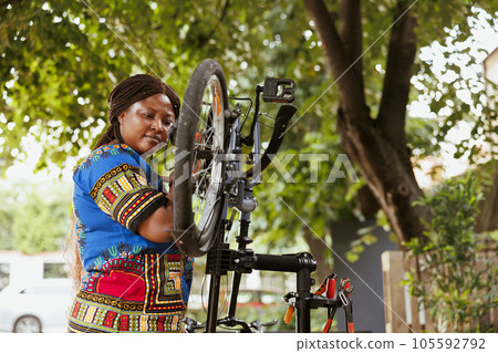 Focused healthy african american woman mending her own bicycle in yard and performing annual outdoor maintenance using expert tools. Female cyclist inspects and repairs bike pedals. 105592792
