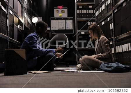 Police officers sitting on floor analyzing crime scene evidence, working at criminology investigation in arhive room. Private detectives checking surveillance footage witch provided vital clue Police officers sitting on floor analyzing crime scene evidence, working at criminology investigation in arhive room. Private detectives checking surveillance footage witch provided vital clue 105592862