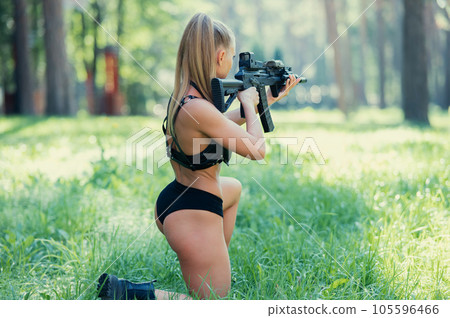 Caucasian woman in bikini sits with machine gun on green grass.  105596466