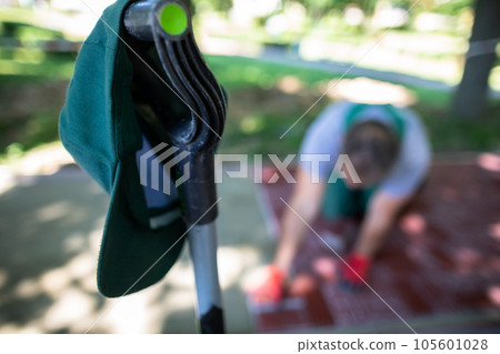 A green baseball cap hangs on the handle of a shovel and in the background a worker is laying paving stones. A green baseball cap hangs on the handle of a shovel and in the background a worker is laying paving stones. 105601028