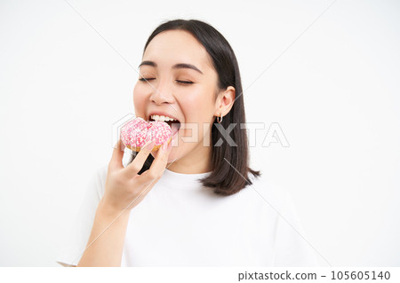 Food and eating out. Young korean woman looks, takes bite of delicious pink glazed doughnut, white background 105605140