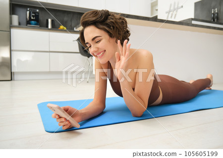 Portrait of happy fitness girl lying on rubber yoga mat, talking on mobile phone, saying hi at smartphone video chat, online training instructor connecting to gym session from home 105605199