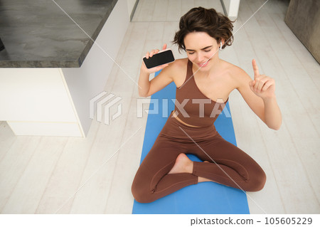 Top view of young active woman, healthy girl stretching, doing workout at home, holding smartphone and listening music in headphones while workout at home, does fitness, pilates yoga on floor mat 105605229
