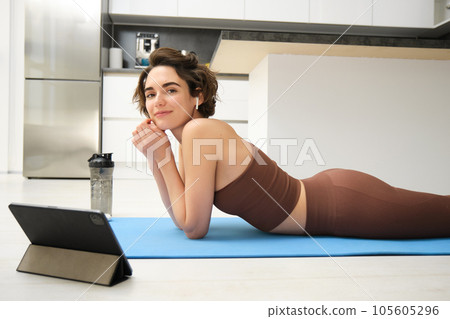 Portrait of beautiful woman in fitness sportswear, lies on floor, rubber yoga mat, uses tablet and wireless headphones, watches online sport workout tutorial, gym classes online from home Portrait of beautiful woman in fitness sportswear, lies on floor, rubber yoga mat, uses tablet and wireless headphones, watches online sport workout tutorial, gym classes online from home 105605296