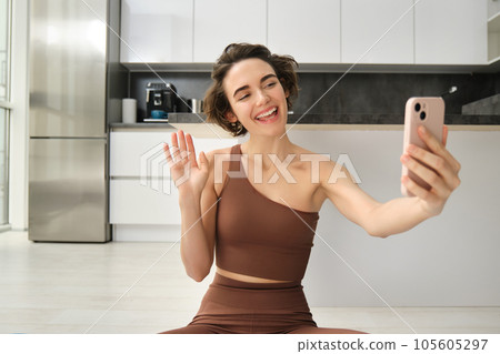Image of young fitness girl blogger, records her workout at home, chats online while doing yoga in bright room. Woman with smartphone, joining remote yoga training via mobile phone Image of young fitness girl blogger, records her workout at home, chats online while doing yoga in bright room. Woman with smartphone, joining remote yoga training via mobile phone 105605297