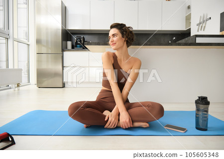 Portrait of fitness girl doing yoga on rubber mat at home, workout indoors in kitchen wearing sportswear, practice minfulness, sitting in lotus pose with water bottle next to her Portrait of fitness girl doing yoga on rubber mat at home, workout indoors in kitchen wearing sportswear, practice minfulness, sitting in lotus pose with water bottle next to her 105605348