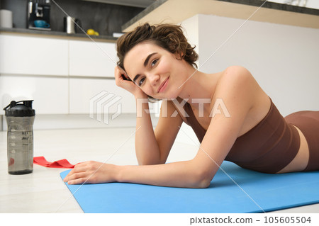 Sport and healthy lifestyle. Portrait of brunette sportswoman doing fitness in activewear, lying on rubber mat on floor, doing workout training in kitchen, drinking water from gym bottle 105605504