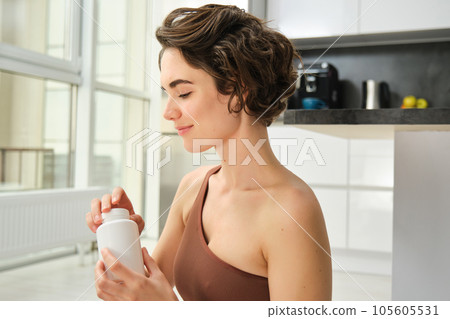 Portrait of young sportswoman, fitness girl takes vitamins, buds for healthy and fit body, workout indoors at home, smiles happily Portrait of young sportswoman, fitness girl takes vitamins, buds for healthy and fit body, workout indoors at home, smiles happily 105605531