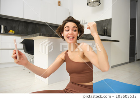Young woman workout at home, rejoicing, using smartphone sport app, online video recordings from fitness instructor, celebrating, sitting on yoga mat on kitchen floor 105605532