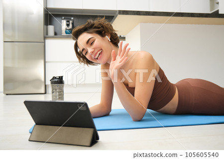 Sport and lifestyle. Young fitness woman, gym instructor waves hand at tablet, joins online video class, yoga and mindfulness workout, training from home indoors Sport and lifestyle. Young fitness woman, gym instructor waves hand at tablet, joins online video class, yoga and mindfulness workout, training from home indoors 105605540