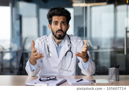 Portrait of a young Indian doctor talking seriously and worriedly to the camera. He sits in the office and communicates with the patient, conducts an online webinar, gestures with his hands. 105607714