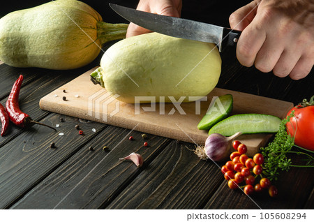 Slicing vegetable marrow with a knife before cooking by the hands of the cook on a wooden cutting board. In cooking, the thick flesh of zucchini is used. 105608294