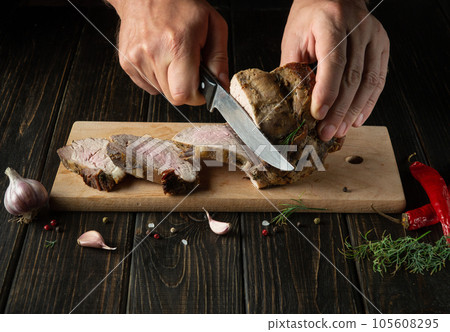 The chef cuts the grilled veal meat into small slices. Work environment on the kitchen table with condiments or spices. Black background with space for recipe or menu. 105608295