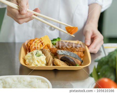 Close-up of a young woman's hand in cooking clothes packing carrots into a lunch box in the kitchen Close-up of a young woman's hand in cooking clothes packing carrots into a lunch box in the kitchen 105610166