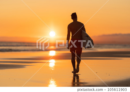 Black silhouette of an attractive man walking on the shore holding a surfboard at orange sunset 105611903