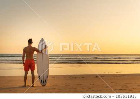 A man standing on the shore holding a surfboard - view from the back 105611915