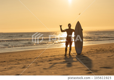 Silhouette of a man standing on the shore holding a surfboard and showing shaka 105611923