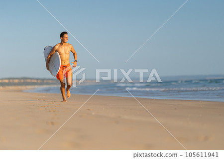 An attractive man with nice athletic body running on the shore holding a surfboard 105611941
