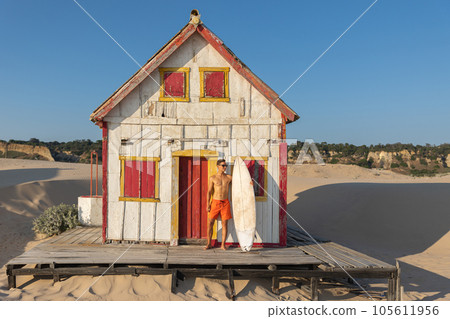 A man with nice athletic body standing at the old seaside house holding a surfboard 105611956
