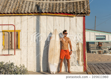 A man with nice athletic body wearing sunglasses standing at the shore house on the beach with his surfboard A man with nice athletic body wearing sunglasses standing at the shore house on the beach with his surfboard 105611960