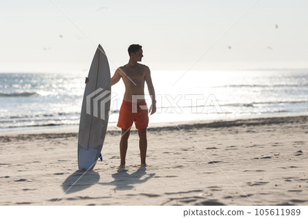 A shirtless man surfer standing on the beach holding a surfboard A shirtless man surfer standing on the beach holding a surfboard 105611989