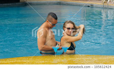 The loving couple hugs and kisses, drinking blue cocktail alcohol liquor in swimming pool at hotel outdoor. Portrait of caucasian man and woman. 105612424