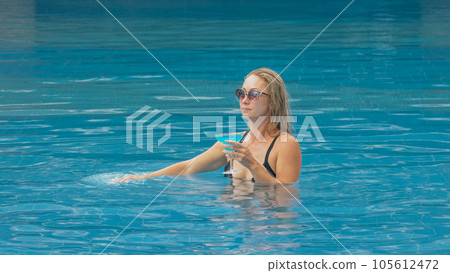 The wide middle shot pretty woman drinking blue cocktail alcohol liquor in swimming pool at hotel. Portrait of sexy girl in pool outdoor. 105612472