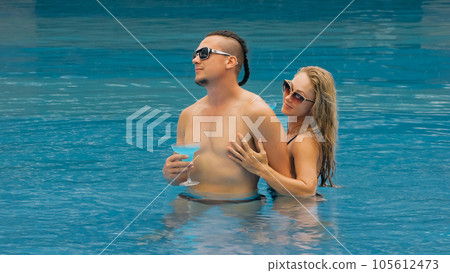 The loving couple hugs and kisses, drinking blue cocktail alcohol liquor in swimming pool at hotel outdoor. Portrait of caucasian man and woman. The loving couple hugs and kisses, drinking blue cocktail alcohol liquor in swimming pool at hotel outdoor. Portrait of caucasian man and woman. 105612473